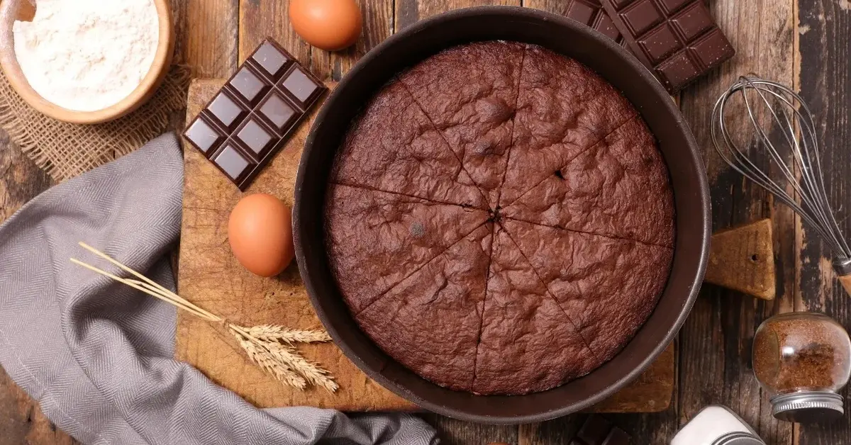 Ciasto czekoladowe w okrągłej formie stojącej na drewnianej desce. Obok leżą jajka, tabliczka czekolady i mąka w misce