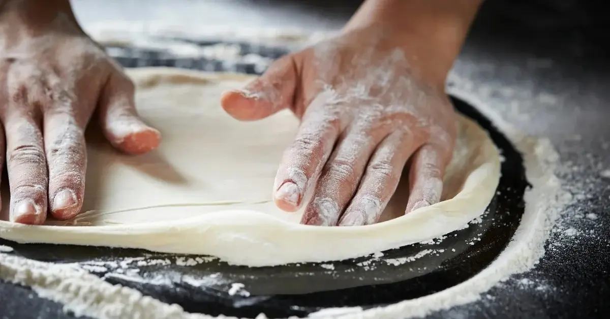 Ciasto na pizzę uformowane na okrągły placek rozkładane dwoma męskimi rękami na podkładzie do pizzy