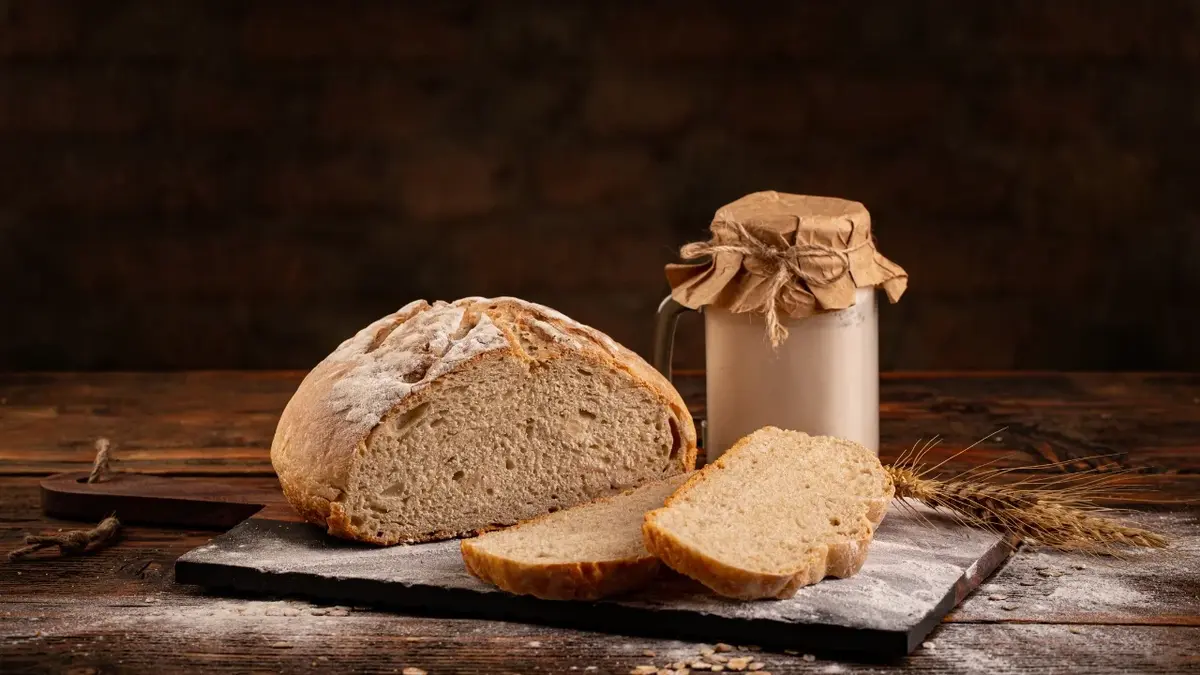 Chrupiący chleb na zakwasie obok dwie kromki ukrojone z bochenka i słoiczek z zakwasem