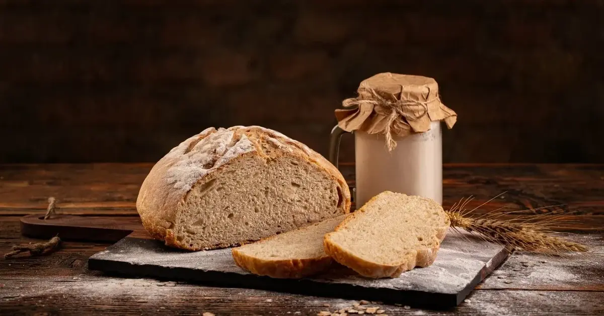 Chrupiący chleb na zakwasie obok dwie kromki ukrojone z bochenka i słoiczek z zakwasem