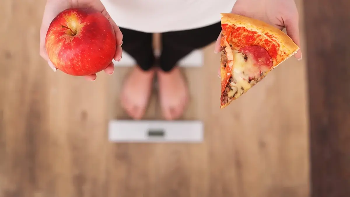 Kobieta stojąca na wadze z jabłkiem w jednej ręce i pizzą w drugiej 