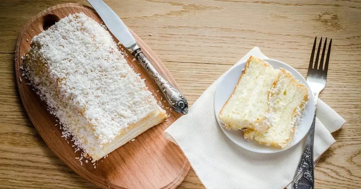 Ciasto anielski puch na drewnianej desce. Obok biała serwetka a na niej biały talerzyk z kawałkiem ciasta. Obok widelec