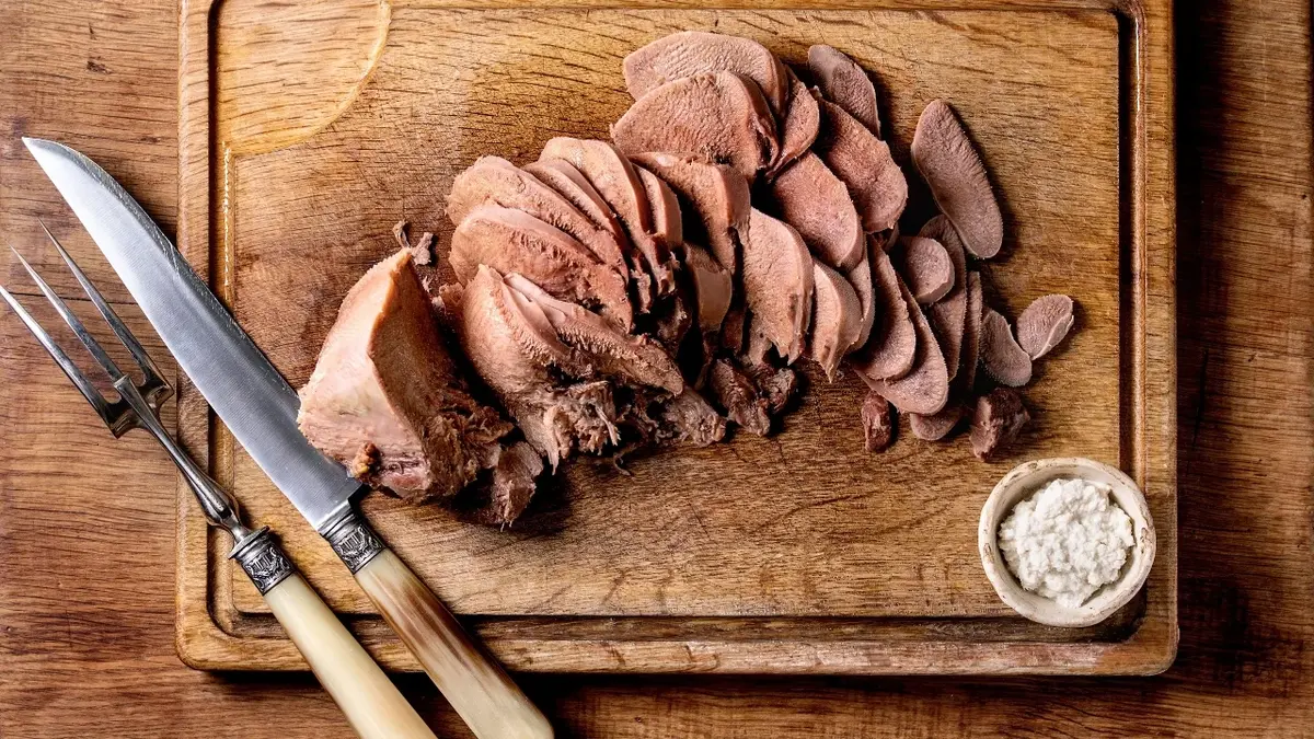 Na drewnianej desce pokrojony na cienkie plasterki kawałek mięsa. Obok srebrny widelec  nóż