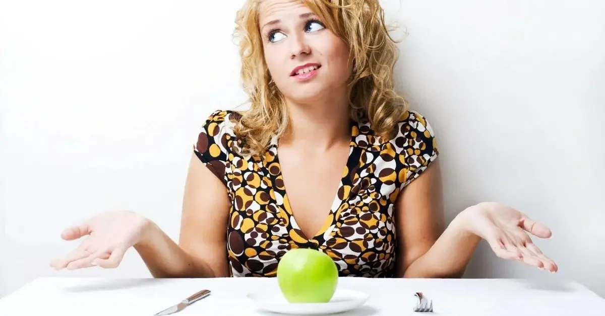 Kobieta o blond włosach, siedząca przy białym stole, rozkłada ręce nad talerzem z zielonym jabłkiem