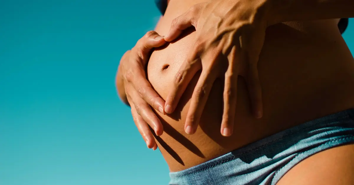 Na niebieskim tle kobiecy brzuch w 3 trymestrze ciąży z dłońmi ułożonymi wokół pępka w kształt serca