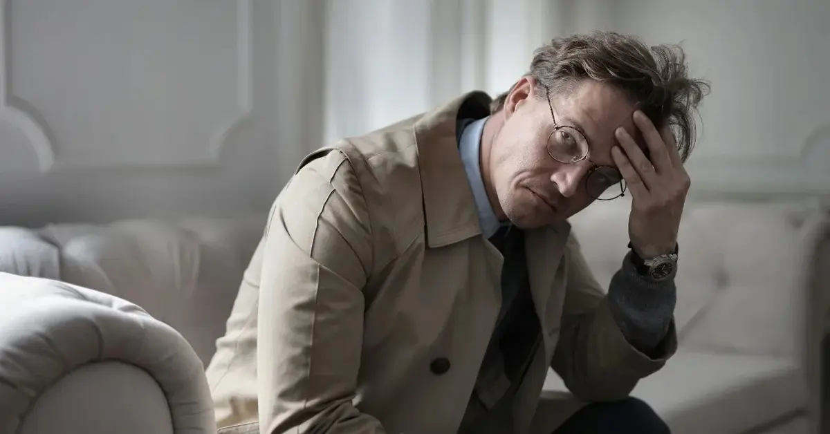 Mężczyzna w płaszczu siedzi na kanapie ze znudzoną miną.